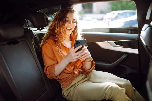 坐在汽车后座上 用智能手机微笑的女人 年轻女人检查邮件 博客在车上 技术概念 — 图库照片