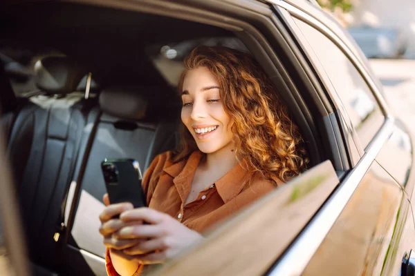 坐在汽车后座上 用智能手机微笑的女人 年轻女人检查邮件 博客在车上 技术概念 — 图库照片