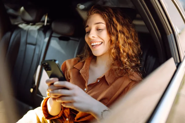 stock image Smiling woman using smartphone while sitting in the back seat of a car. Young woman checks mail, texts, blogs in the car. Business, technology concept.