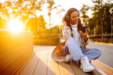 Şehirde akıllı telefon ve kulaklık kullanarak mutlu gülümseyen genç bir kadın müzik dinliyor, sesli yayın yapıyor. Sosyal ağlarda iletişim. Yaşam biçimi. 