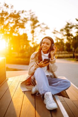 Şehirde akıllı telefon ve kulaklık kullanarak mutlu gülümseyen genç bir kadın müzik dinliyor, sesli yayın yapıyor. Sosyal ağlarda iletişim. Yaşam biçimi. 