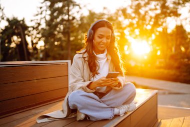 Şehirde akıllı telefon ve kulaklık kullanarak mutlu gülümseyen genç bir kadın müzik dinliyor, sesli yayın yapıyor. Sosyal ağlarda iletişim. Yaşam biçimi. 