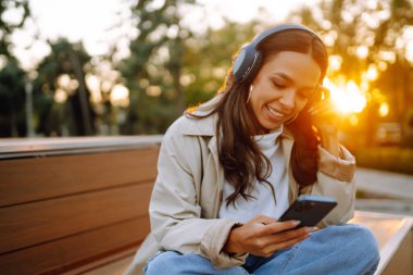 Şehirde akıllı telefon ve kulaklık kullanarak mutlu gülümseyen genç bir kadın müzik dinliyor, sesli yayın yapıyor. Sosyal ağlarda iletişim. Yaşam biçimi. 