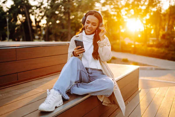 Şehirde akıllı telefon ve kulaklık kullanarak mutlu gülümseyen genç bir kadın müzik dinliyor, sesli yayın yapıyor. Sosyal ağlarda iletişim. Yaşam biçimi. 