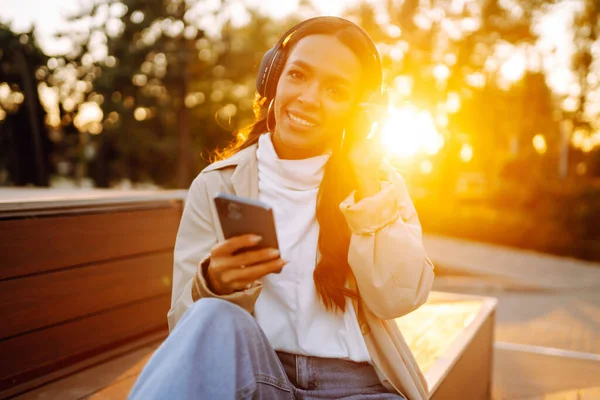 Şehirde akıllı telefon ve kulaklık kullanarak mutlu gülümseyen genç bir kadın müzik dinliyor, sesli yayın yapıyor. Sosyal ağlarda iletişim. Yaşam biçimi. 