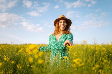 Parlak elbiseli ve zarif şapkalı güzel bir kadın yakut tohumu tarlasında eğleniyor. Gülümseyen dişi turist çiçek tarlasında yürüyor, sarı çiçeklere dokunuyor. Doğa, dinlenme. Yaz manzarası.