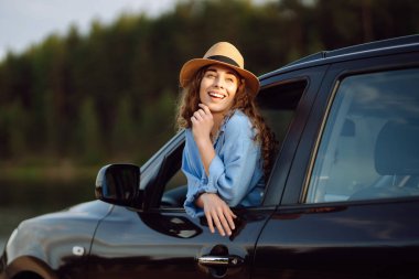 Mutlu kadın dinleniyor ve arabanın camından sarkan günbatımının tadını çıkarıyor. Şapkalı kıvırcık turist kadın yaz gezisinde eğleniyor ve özgürlüğünü hissediyor. Aktif yaşam tarzı, doğa, turizm.