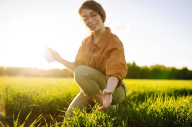 Buğday tarlasında deneyimli çiftçi kadın buğday filizlerini inceliyor. Gülümseyen çiftçi elinde dijital tabletle buğday tarlasının kalitesini kontrol ediyor. Buğday tarlasının ilerleyişini kontrol ediyorum. Tarım kavramı.