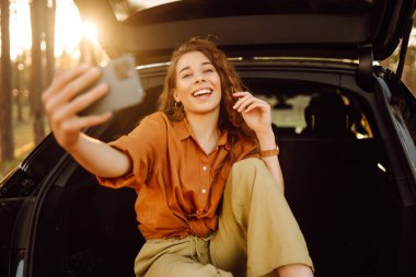 Kadın arabanın bagajında telefonla dinleniyor. Video aracılığıyla mutlu turist sohbetleri ya da arabanın arka koltuğunda selfie çekmeler. Arabayla seyahat etme kavramı, aktif yaşam tarzı.