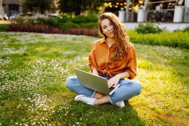 Parktaki yeşil çimenlikteki genç kıvırcık kadın elinde dizüstü bilgisayarla. Gülümseyen serbest çalışan, dışarıda çalışan, güzel havanın tadını çıkaran biri. Serbest çalışma konsepti. Çevrimiçi eğitim.