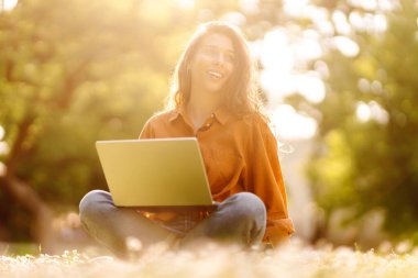 Parktaki yeşil çimenlikteki genç kıvırcık kadın elinde dizüstü bilgisayarla. Gülümseyen serbest çalışan, dışarıda çalışan, güzel havanın tadını çıkaran biri. Serbest çalışma konsepti. Çevrimiçi eğitim.