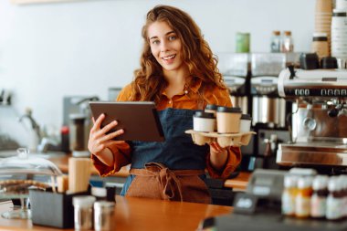 Kıvırcık saçlı ve elinde tablet olan bir kahve dükkanının yakışıklı genç müdürü kahve dükkanında paket kahve veriyor. Paket yemek. İş konsepti.