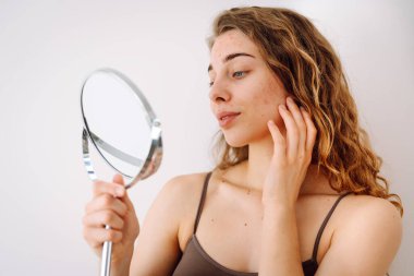Deri sorunu. Kıvırcık kadın parlak bir odada aynaya bakarken yüzündeki kırmızı iltihaplara dokunuyor. Yüz derisi sorunları. Güzellik bakımı. Alerjik reaksiyon.