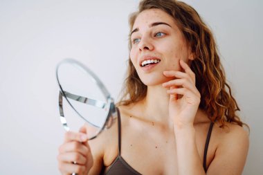 Deri sorunu. Kıvırcık kadın parlak bir odada aynaya bakarken yüzündeki kırmızı iltihaplara dokunuyor. Yüz derisi sorunları. Güzellik bakımı. Alerjik reaksiyon.