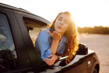 Güzel genç bir kadın yolculuk sırasında araba penceresinden gün batımını izlemeyi sever. Araba yolculuğu, aktif yaşam tarzı konsepti, gün batımı ışığı..