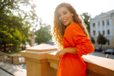 Parlak elbiseli mutlu genç manken şehir manzarasına karşı poz veriyor. Kıvırcık kadın şehrin güzel güneşli havasının tadını çıkarıyor. Yaşam tarzı konsepti. Sokaktaki fotoğraf..