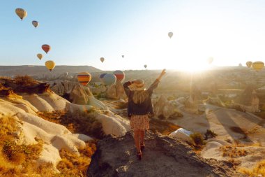 Şapkalı çekici genç bir kadın sırtı dağda duruyor ve uçan sıcak hava balonlarının keyfini çıkarıyor. Anın tadını çıkar. Özgürlük kavramı, aktif yaşam tarzı. Ünlü turizm bölgesi Kapadokya.