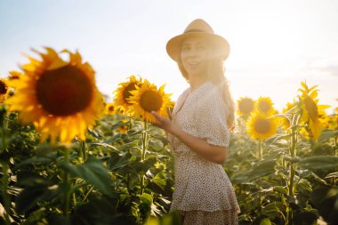 Güneşli, güzel, mutlu bir genç kadının fotoğrafı, sıcak havanın tadını çıkarırken, çiçek açan ayçiçeği tarlasında yürürken. Kadın ayçiçeği tarlasında elbise ve şapkayla poz veriyor. Anın tadını çıkar. Etkin yaşam biçimi.