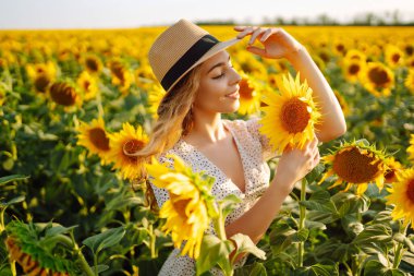 Güneşli, güzel, mutlu bir genç kadının fotoğrafı, sıcak havanın tadını çıkarırken, çiçek açan ayçiçeği tarlasında yürürken. Kadın ayçiçeği tarlasında elbise ve şapkayla poz veriyor. Anın tadını çıkar. Etkin yaşam biçimi.