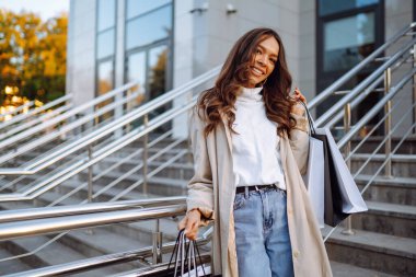 Alışveriş merkezinin yakınındaki alışveriş torbalarıyla Avrupalı çekici kahverengi saçlı bir kadının portresi. Güneşli bir günde alışveriş çantalı şık genç bir kadın şehrin sokaklarında yürüyor. Tüketim, alışveriş