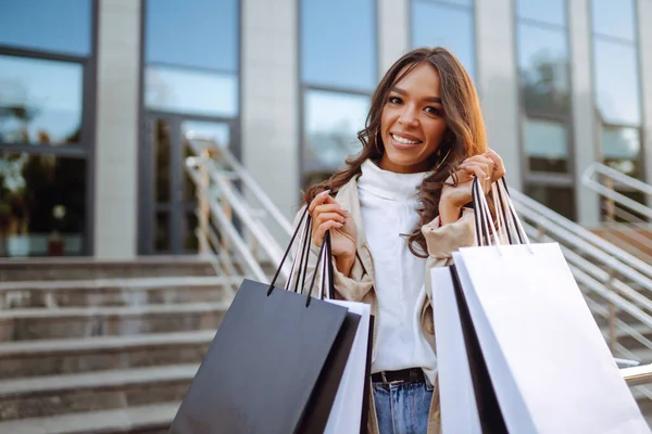 一个有魅力的棕色头发的女人的肖像 她来自欧洲 在购物中心附近带着购物袋 一个阳光灿烂的日子 一个带着购物袋的时髦年轻女子走过城市的街道 消费主义 — 图库照片
