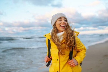 Sarı ceketli genç bir turist deniz kenarında yürüyor, gün batımında deniz manzarasının tadını çıkarıyor. Seyahat, turizm konsepti. Etkin yaşam biçimi.