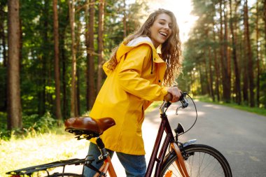 Sarı ceketli, kıvırcık saçlı gülümseyen kadın güneşli bir parkta bisiklet sürüyor. Turist kadın sonbahar doğasını sever. Doğa kavramı ve aktif yaşam tarzı.
