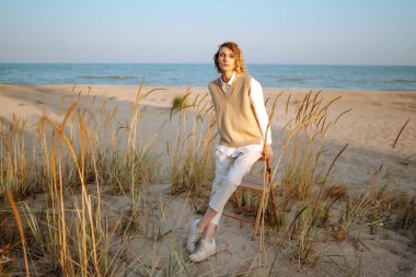 Şık elbiseli, rüzgarlı saçlı ince manken sahildeki yabani otların arasında poz veriyor. Rüzgarlı sahilde duran güzel bir kadın. Özgürlük için. Moda anlayışı, rahatlama, dinlenme.
