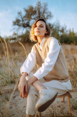 Şık elbiseli, rüzgarlı saçlı ince manken sahildeki yabani otların arasında poz veriyor. Rüzgarlı sahilde duran güzel bir kadın. Özgürlük için. Moda anlayışı, rahatlama, dinlenme.