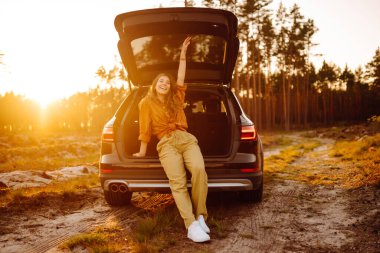 Gülümseyen güzel kadın gün batımında bir arabanın bagajında oturuyor ve manzaranın tadını çıkarıyor. Seyahat, tatil, yaşam tarzı konsepti.