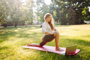 Güzel bir kadın sabahları ya da gün batımında parkta bilgisayarla yoga yapıyor. Sporcu kadın yeşil bir çimenlikte jimnastik halısı üzerinde gerinmekle meşgul. Fitness.