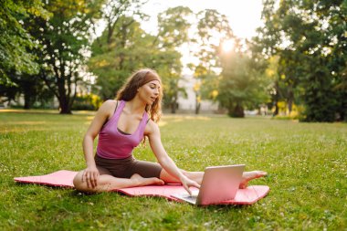 Güzel bir kadın sabahları ya da gün batımında parkta bilgisayarla yoga yapıyor. Sporcu kadın yeşil bir çimenlikte jimnastik halısı üzerinde gerinmekle meşgul. Fitness.