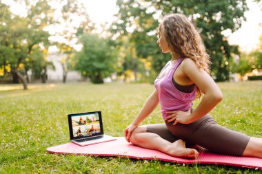 Güzel bir kadın sabahları ya da gün batımında parkta bilgisayarla yoga yapıyor. Sporcu kadın yeşil bir çimenlikte jimnastik halısı üzerinde gerinmekle meşgul. Fitness.
