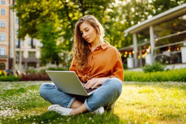 Yeşil bir çayırda dizüstü bilgisayarla oturan genç ve güzel bir kadının portresi. Eğitim çevrimiçi. Dışarı koşan mutlu kadın.