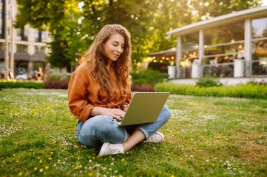 Yeşil bir çayırda dizüstü bilgisayarla oturan genç ve güzel bir kadının portresi. Eğitim çevrimiçi. Dışarı koşan mutlu kadın.