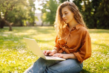 Yeşil bir çayırda dizüstü bilgisayarla oturan genç ve güzel bir kadının portresi. Eğitim çevrimiçi. Dışarı koşan mutlu kadın.