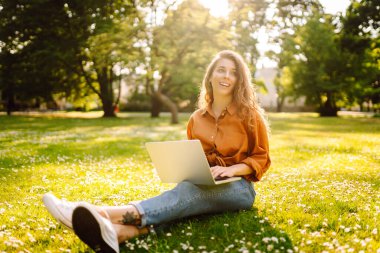 Yeşil bir çayırda dizüstü bilgisayarla oturan genç ve güzel bir kadının portresi. Eğitim çevrimiçi. Dışarı koşan mutlu kadın.