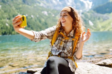 Dağın tepesinde kıvırcık saçlı, güzel bir turist kadın. Aktif kadın telefonla video bağlantısıyla konuşuyor, selfie çekiyor. Etkin bir yaşam biçimi kavramı.
