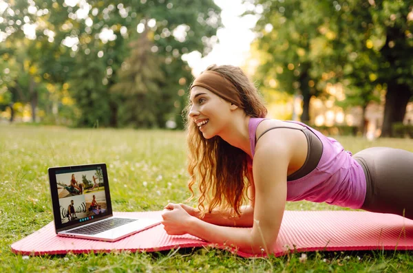 Beautiful woman practices yoga with a laptop via video link in the morning or in the park at sunset. Sportswoman is engaged in stretching on a gymnastic carpet on a green lawn. Fitness.