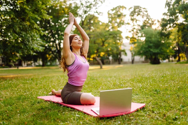 Beautiful woman practices yoga with a laptop via video link in the morning or in the park at sunset. Sportswoman is engaged in stretching on a gymnastic carpet on a green lawn. Fitness.
