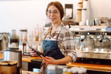Başarılı küçük işletme sahibi dijital tabletle kahve dükkanının arkasında duruyor ve düzeni sağlıyor. Güzel kadın baristanın portresi. Satıcı-girişimci iş konsepti