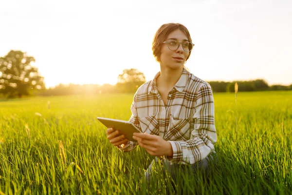 Young farmer woman with digital tablet evaluates shoots, green wheat sprouts in the field. Farm work. Technology of modern agriculture. Harvesting. Concept of ecology, gardening.