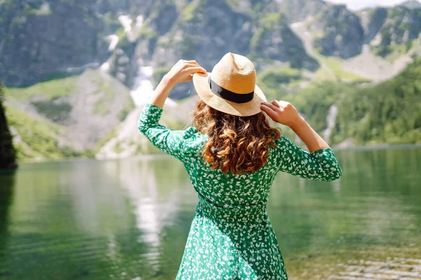 Parlak yeşil elbiseli ve şapkalı genç turist kadın dağların ve turkuaz dağ gölünün arka planında. Mutlu kadın dağ manzarasını seviyor. Turizm kavramı, tatil.