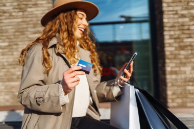 Şapkalı, alışveriş çantalı, kredi kartlı, telefonlu güzel bir kadın. Kıvırcık kadın kredi kartıyla alışveriş yapıyor, güneşli bir sokakta yürüyor. E-ticaret, internet bankacılığı. Yaşam tarzı kavramı.