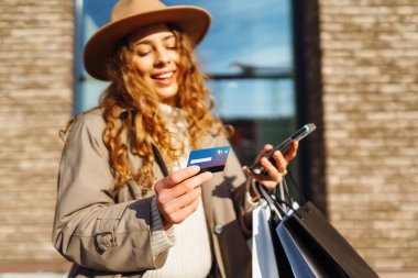 Şapkalı, alışveriş çantalı, kredi kartlı, telefonlu güzel bir kadın. Kıvırcık kadın kredi kartıyla alışveriş yapıyor, güneşli bir sokakta yürüyor. E-ticaret, internet bankacılığı. Yaşam tarzı kavramı.