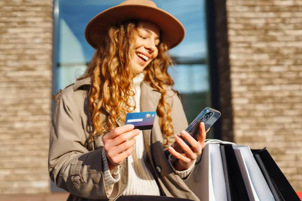 Şapkalı, alışveriş çantalı, kredi kartlı, telefonlu güzel bir kadın. Kıvırcık kadın kredi kartıyla alışveriş yapıyor, güneşli bir sokakta yürüyor. E-ticaret, internet bankacılığı. Yaşam tarzı kavramı.