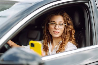 Şık bir kadın araba kullanıyor ve cep telefonu kullanıyor. Şehri dolaştım, park için yas tuttum. İş, teknoloji kavramı.