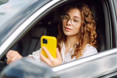 Şık bir kadın araba kullanıyor ve cep telefonu kullanıyor. Şehri dolaştım, park için yas tuttum. İş, teknoloji kavramı.