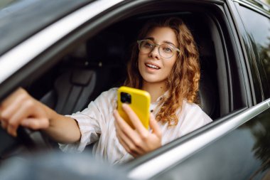Şık bir kadın araba kullanıyor ve cep telefonu kullanıyor. Şehri dolaştım, park için yas tuttum. İş, teknoloji kavramı.