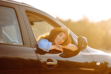 Arabanın penceresinden gülümseyen kadın doğanın tadını çıkarıyor. Arabayla seyahat ederken özgürlüğü hissediyorsunuz. Araba yolculuğu, gün batımı. Etkin yaşam biçimi.